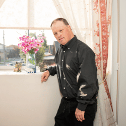 Disabled man with down syndrome at home leaning against window sill and smiling at camera