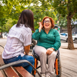 Disabled woman in wheelchair talking with friend sitting on park bench