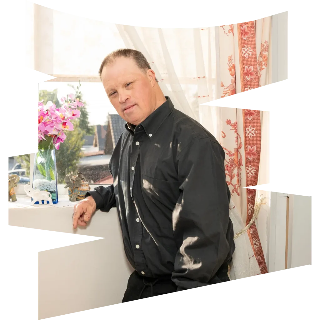 Disabled man with down syndrome at home leaning against window sill and smiling at camera